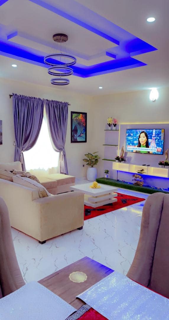 A modern living area is characterized by soft beige furnishings, framed art on light-colored walls, and blue accent lighting. A wall-mounted TV is positioned above a stylish media unit, which hosts decorative items. Large curtains frame the window, allowing natural light to filter in.