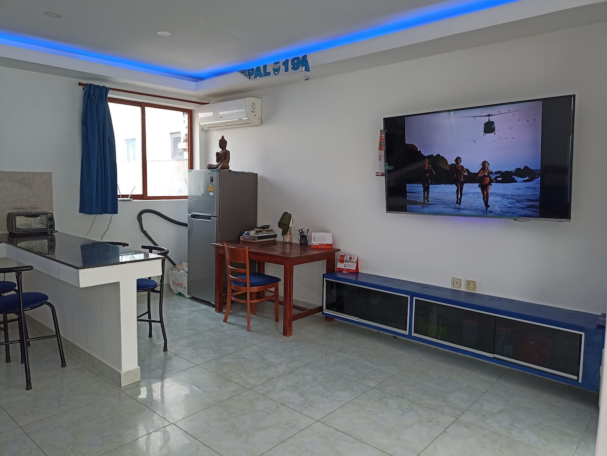 A spacious living area is presented, featuring a large smart TV mounted on the wall opposite a wooden dining table. A kitchen area with a peninsula is visible, alongside a large refrigerator. Natural light enters through a window adorned with blue curtains.