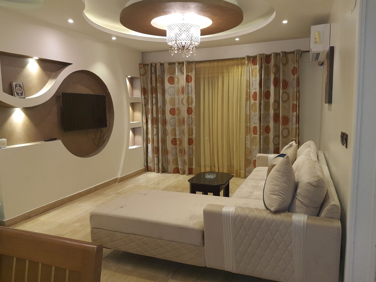 A modern living area is highlighted by a sectional sofa and a minimalist coffee table. Soft lighting is provided by a chandelier, while large windows adorned with patterned curtains offer natural light. A wall-mounted television is situated opposite the seating area.