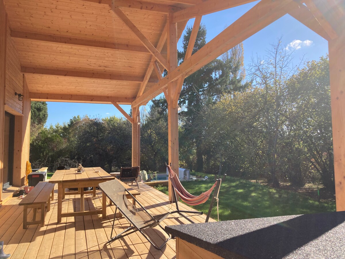A spacious outdoor terrace is highlighted by wooden beams and a sunlit atmosphere, featuring a large table for gatherings, lounge chairs, and a hammock. The surrounding greenery creates a serene backdrop, providing a comfortable area for relaxation and enjoyment.