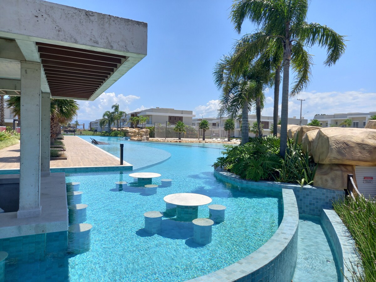 A serene pool area is depicted, featuring a winding design surrounded by lush greenery and palm trees. Several round tables are partially submerged in the water, providing a unique seating arrangement. The clear blue water reflects the bright sky, creating a tranquil atmosphere.