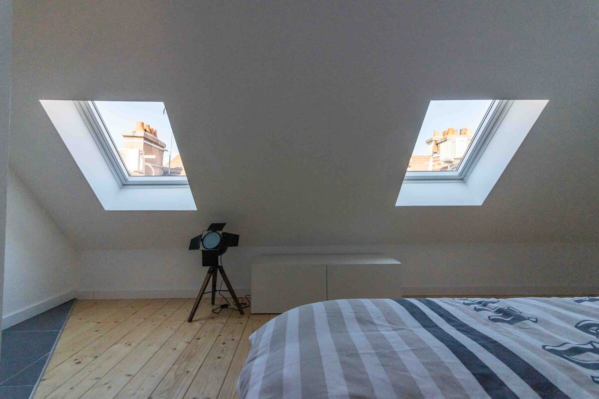 A cozy bedroom is displayed with slanted ceilings and two skylights that provide natural light and views of the sky. The light wood flooring complements the space, while a modern black floor lamp stands nearby, enhancing the room's contemporary design.