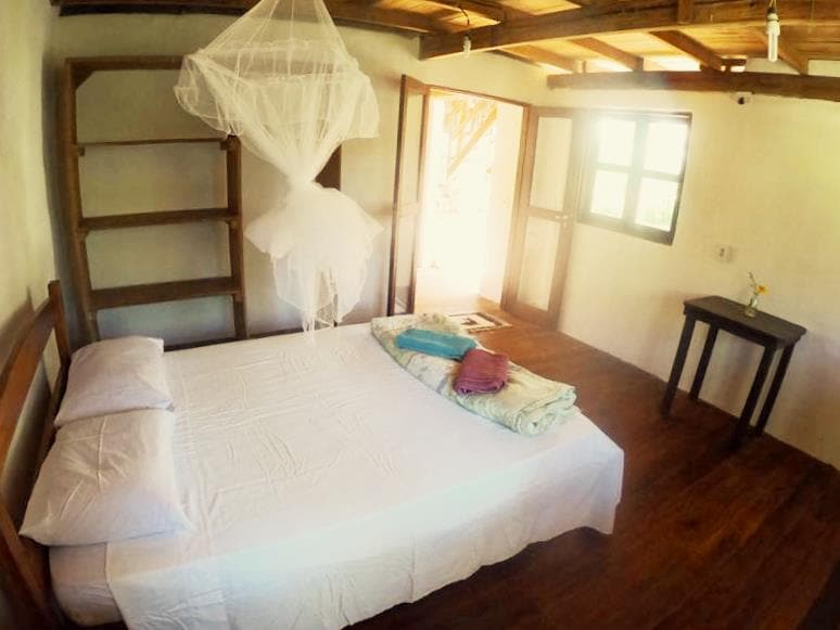 A spacious bedroom is showcased with a double bed dressed in fresh white linens. A mosquito net hangs gracefully above the bed. Wooden shelves are visible, alongside a small table near the window, which allows natural light to fill the room.