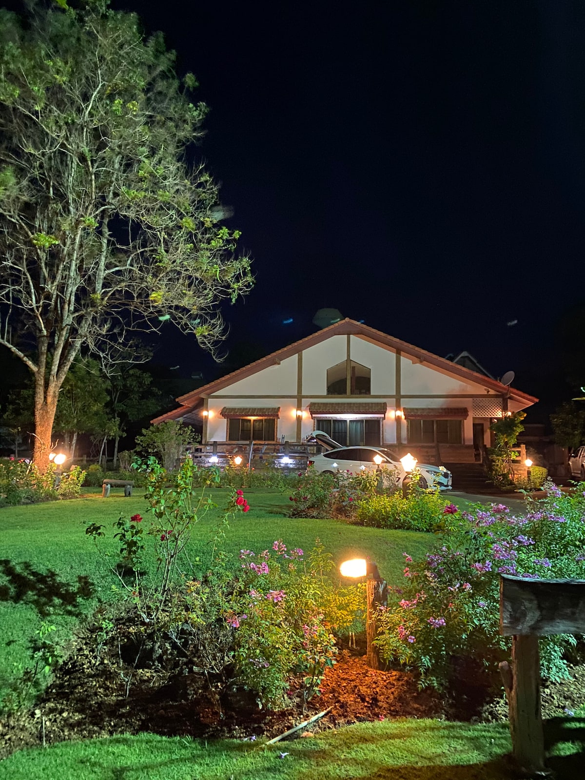The exterior of the house is illuminated at night, showcasing its warm lighting. A well-manicured garden features various flowers, and a large tree adds greenery to the tranquil setting. The spacious lawn invites relaxation and is bordered by decorative lighting.