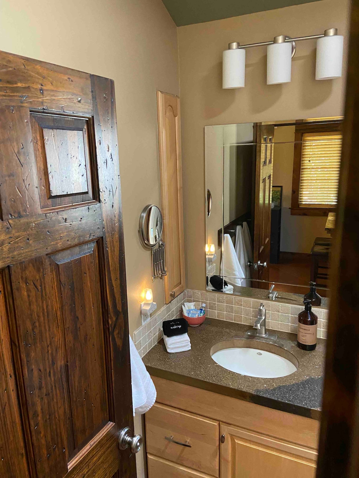 A bathroom features natural wood accents and a large mirror above a countertop. A round sink is set into the stone-like surface. Soft lighting illuminates the area, with hand towels and personal amenities neatly arranged nearby.