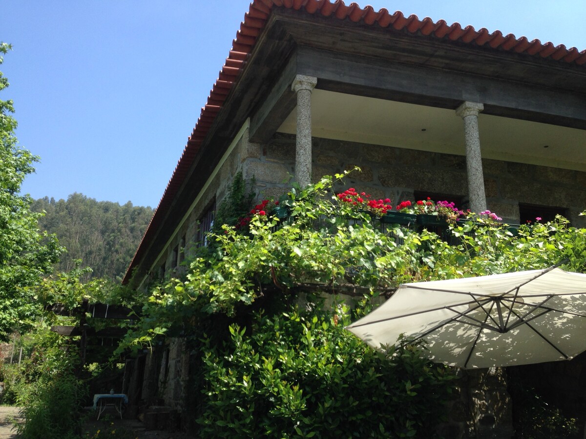 The exterior of the stone house features a traditional red-tiled roof and elegant columns. Lush greenery, including climbing vines and blooming flowers, adorn the balcony, creating a vibrant and inviting atmosphere. A shaded seating area with an umbrella is visible in the foreground.