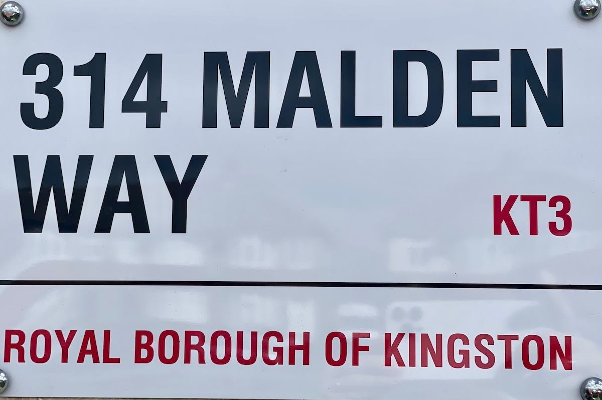 A street sign displaying '314 Malden Way' in bold black letters. The postal code 'KT3' is noted above, while the phrase 'Royal Borough of Kingston' is highlighted in red. The background features a blurred view of nearby buildings.