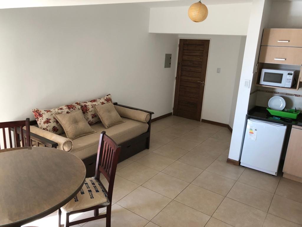 A living area features a sofa bed adorned with decorative pillows, complemented by a round dining table with four chairs. A kitchenette is visible with a microwave and refrigerator, while a wooden door leads to another room.