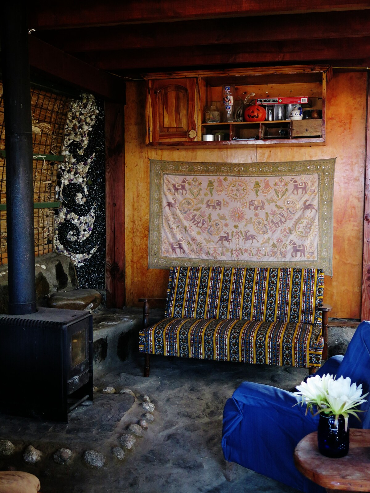 A rustic living area features a patterned sofa against a wooden wall, complemented by a decorative tapestry above. A wood-burning stove adds warmth to the space, while decorative elements made of natural materials enhance the earthy aesthetic.