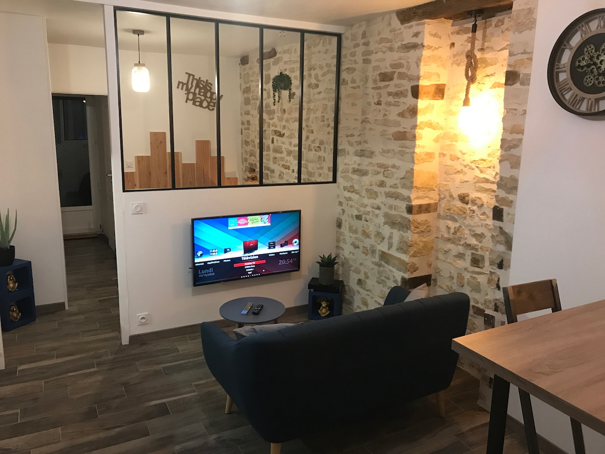 A cozy living area features a dark blue sofa facing a wall-mounted TV. Textured stone walls and warm lighting create a welcoming environment. A wooden dining table and decorative plants are positioned nearby, enhancing the spacious ambiance within the apartment.