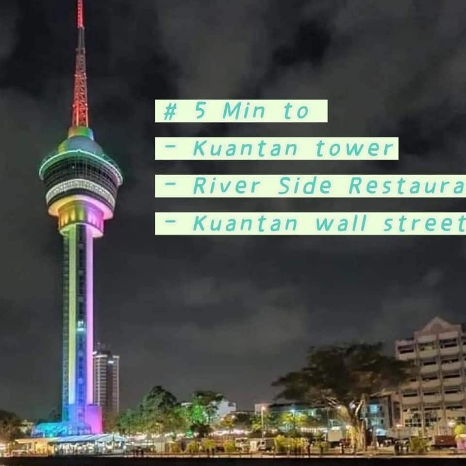 The Kuantan Tower is depicted at night, showcasing vibrant lights that illuminate its structure. Surrounding buildings and trees are visible, contributing to the urban landscape. Clear signage indicates the proximity to local attractions, emphasizing accessibility to dining and entertainment options.