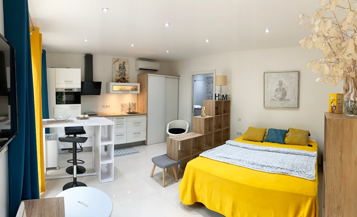 A well-appointed studio space features a vibrant yellow bed and a modern kitchenette, complemented by a comfortable dining area. Ample natural light highlights the contemporary furnishings and decor, while built-in storage provides functionality. A television is mounted on the wall, enhancing entertainment options.