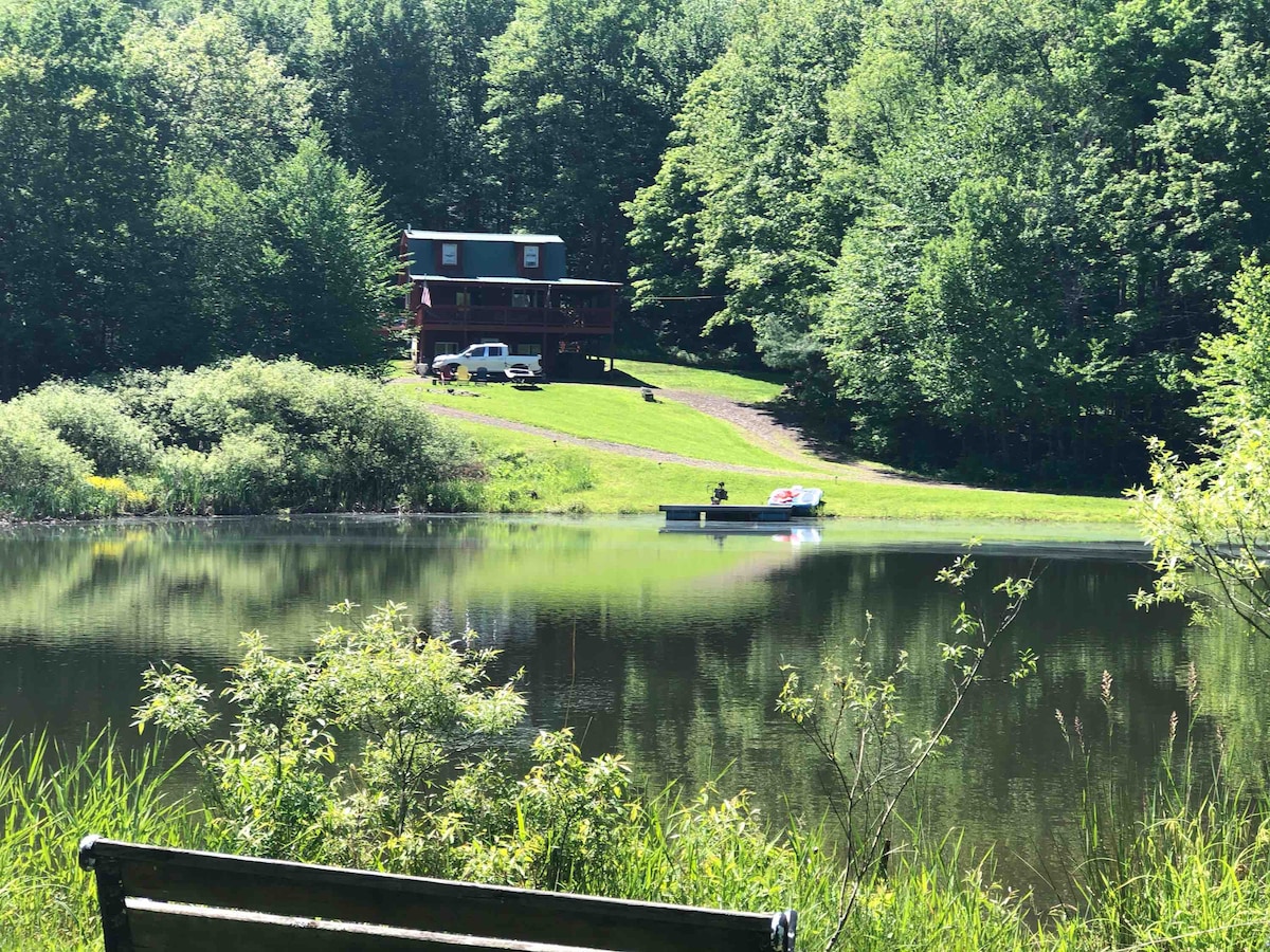 A serene landscape is captured, featuring a tranquil pond bordered by greenery. A cozy cabin is visible in the background, nestled among trees with a green lawn leading up to it. A small boat is stationed on the water, enhancing the peaceful setting.