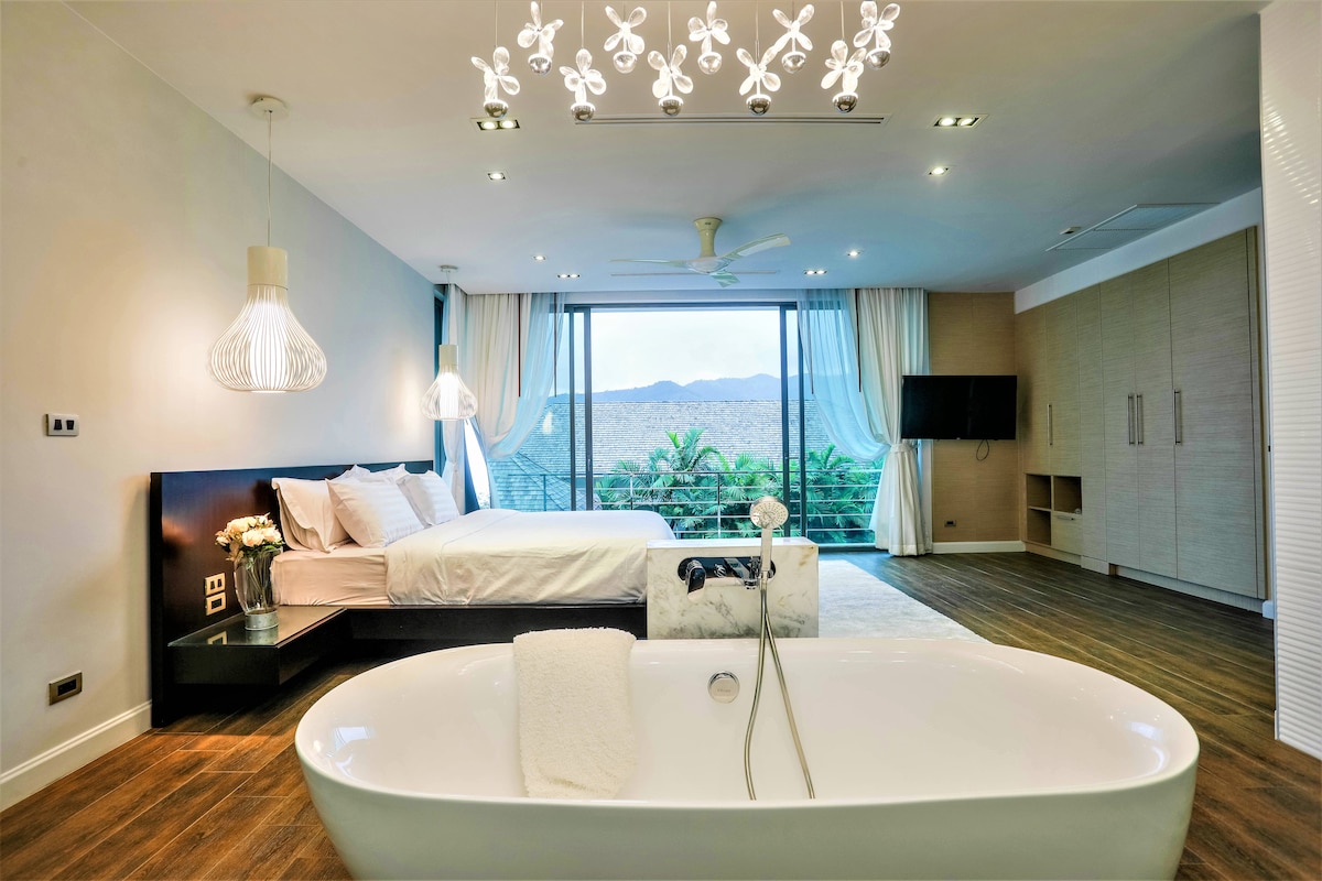 An airy bedroom features a spacious bed with crisp linens, illuminated by modern pendant lights. A freestanding bathtub is positioned in the foreground, while floor-to-ceiling windows provide a view of lush greenery beyond. Neutral tones and wooden flooring enhance the room's modern aesthetic.