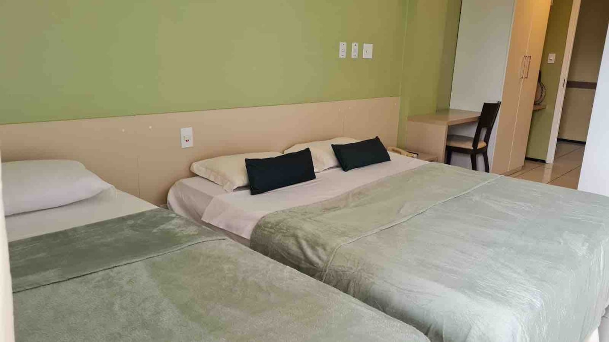 The sleeping area features two spacious beds covered with soft green bedspreads. Neutral-colored pillows are placed at the head of each bed. A simple wooden chair and desk are visible in the background, and light-colored walls create a calm atmosphere.