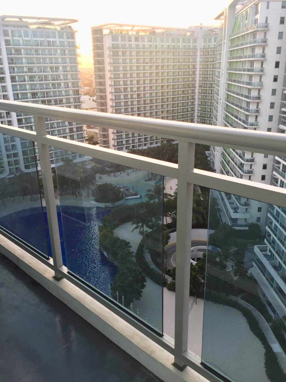 A balcony railing made of glass provides a clear view of the surrounding buildings and a blue wave pool below. The sunset casts a warm glow over the scene, highlighting the lush greenery and pathways of the complex.