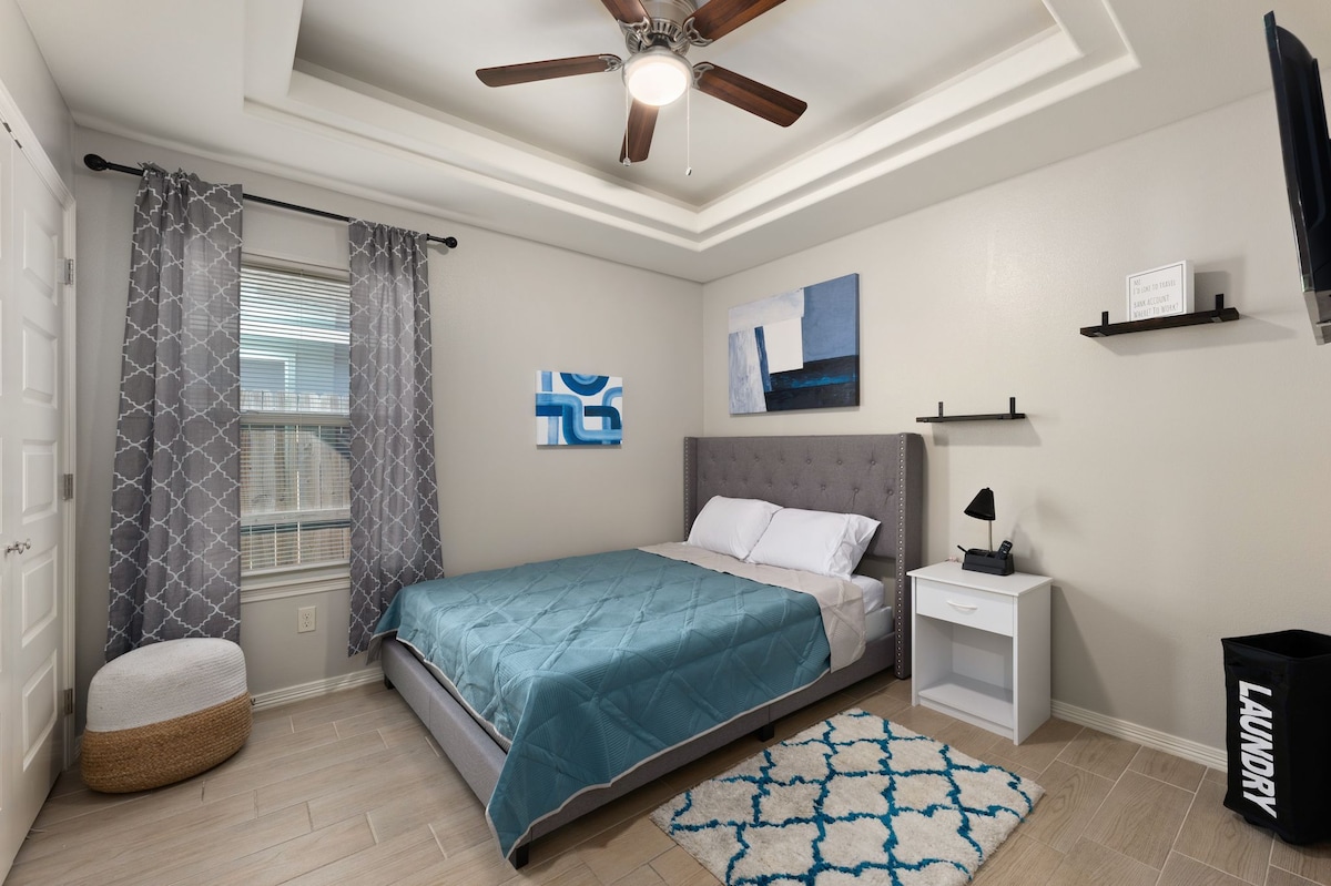 A bedroom features a spacious bed covered with a bright blue quilt. Gray curtains frame a window, allowing natural light to illuminate the room. A light fixture with wooden blades hangs above, while a small bedside table with a lamp sits beside the bed. Decorative artwork and a shelving unit add visual interest.