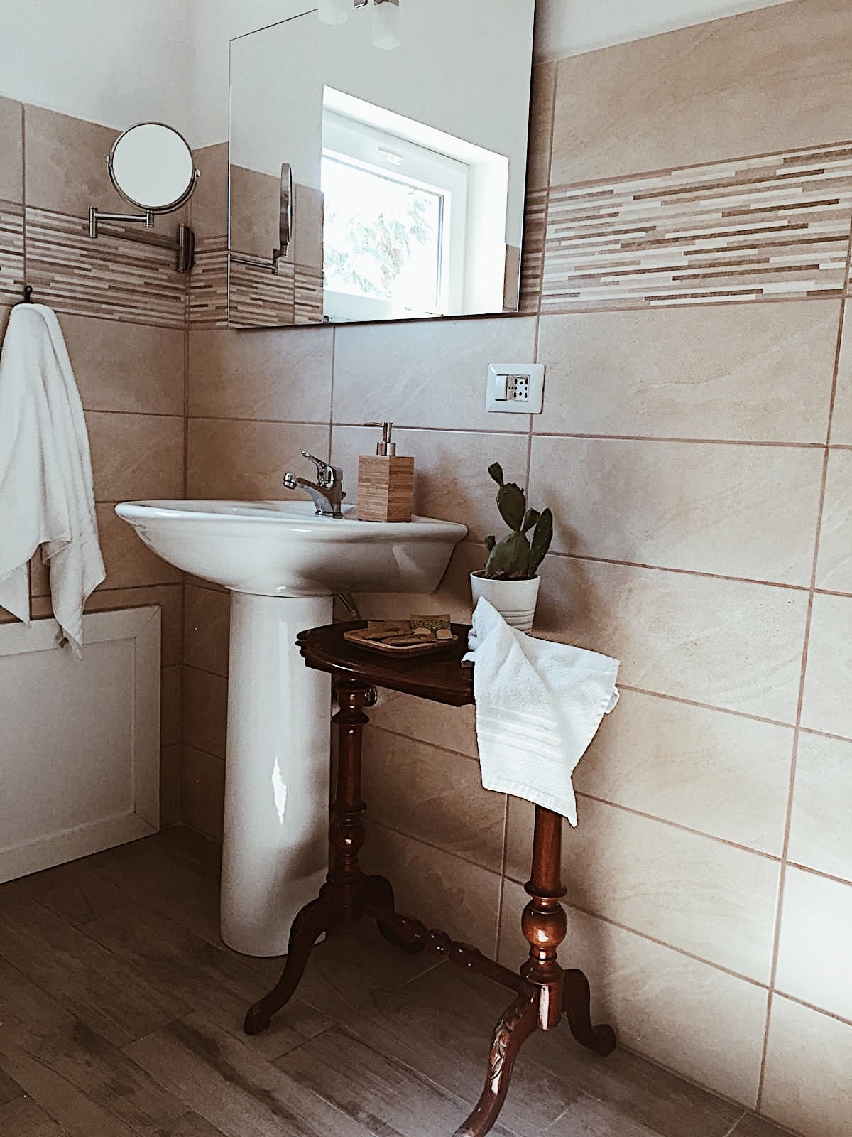 The bathroom features a white pedestal sink beneath a rectangular mirror. Light beige tiles cover the walls, complemented by decorative strips. A small wooden table displays toiletries and a potted cactus. A towel hangs nearby, adding to the functional and simple design.