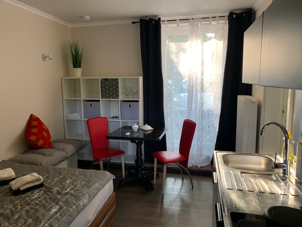 A compact room showcases a spacious bed with neutral bedding. A small dining table with two red chairs is positioned near a window with sheer curtains. A kitchenette is visible, along with a minimalist shelving unit and a cozy seating area.