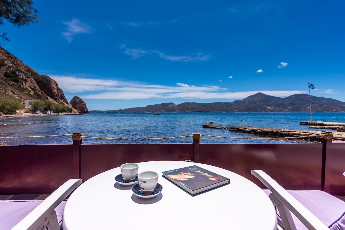 A circular table is set for two, featuring two cups and a magazine. The backdrop reveals a serene coastline with calm waters and distant hills under a clear blue sky. A flag can be seen near the water's edge, accompanied by wooden seating areas along the shore.