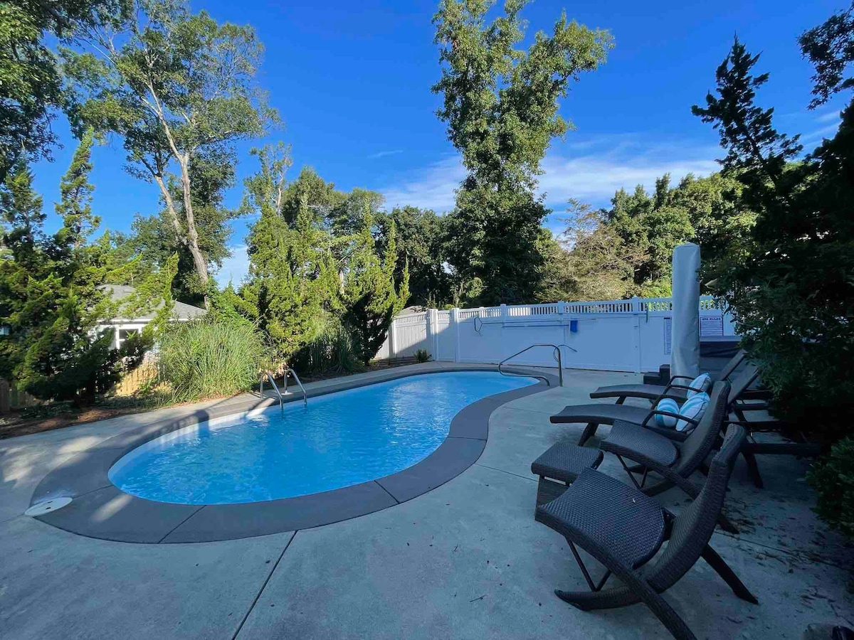 A private pool is surrounded by a concrete patio, featuring multiple loungers with thick cushions. Lush greenery borders the area, providing a serene backdrop against a clear blue sky.