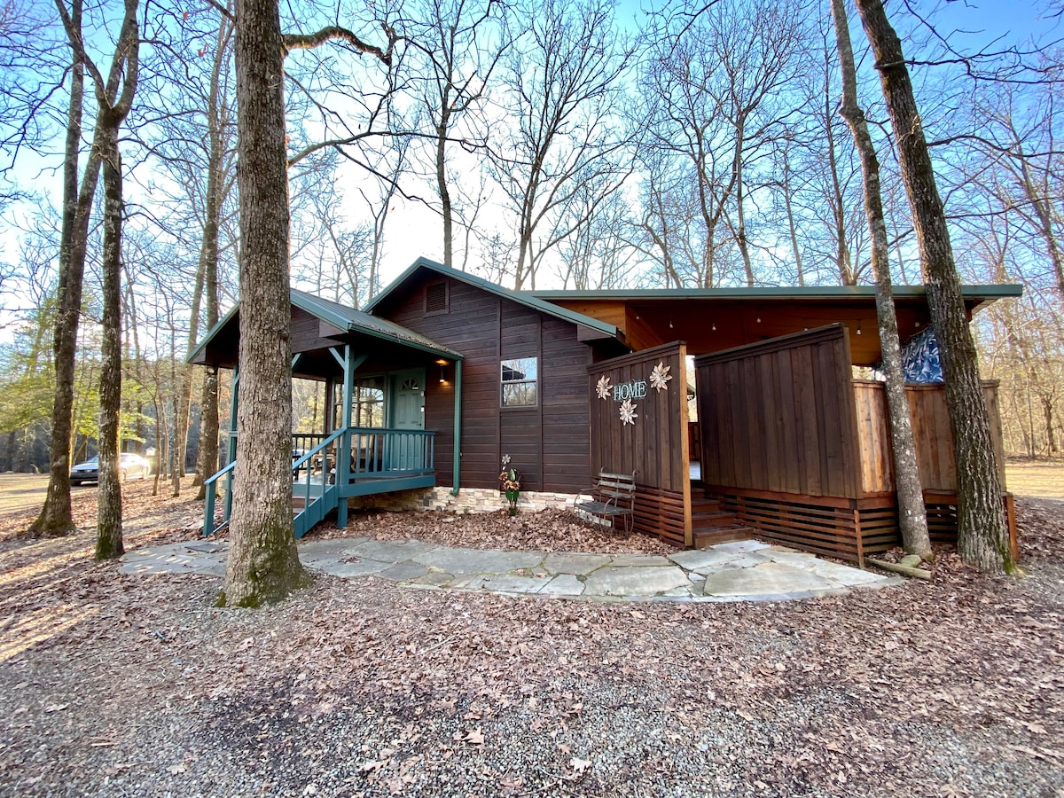 The cabin exterior is framed by tall trees, showcasing a wooden facade with a welcoming entrance. A stone walkway leads to a covered porch, providing an inviting outdoor space amid the natural surroundings. An outdoor privacy screen is visible on the side.
