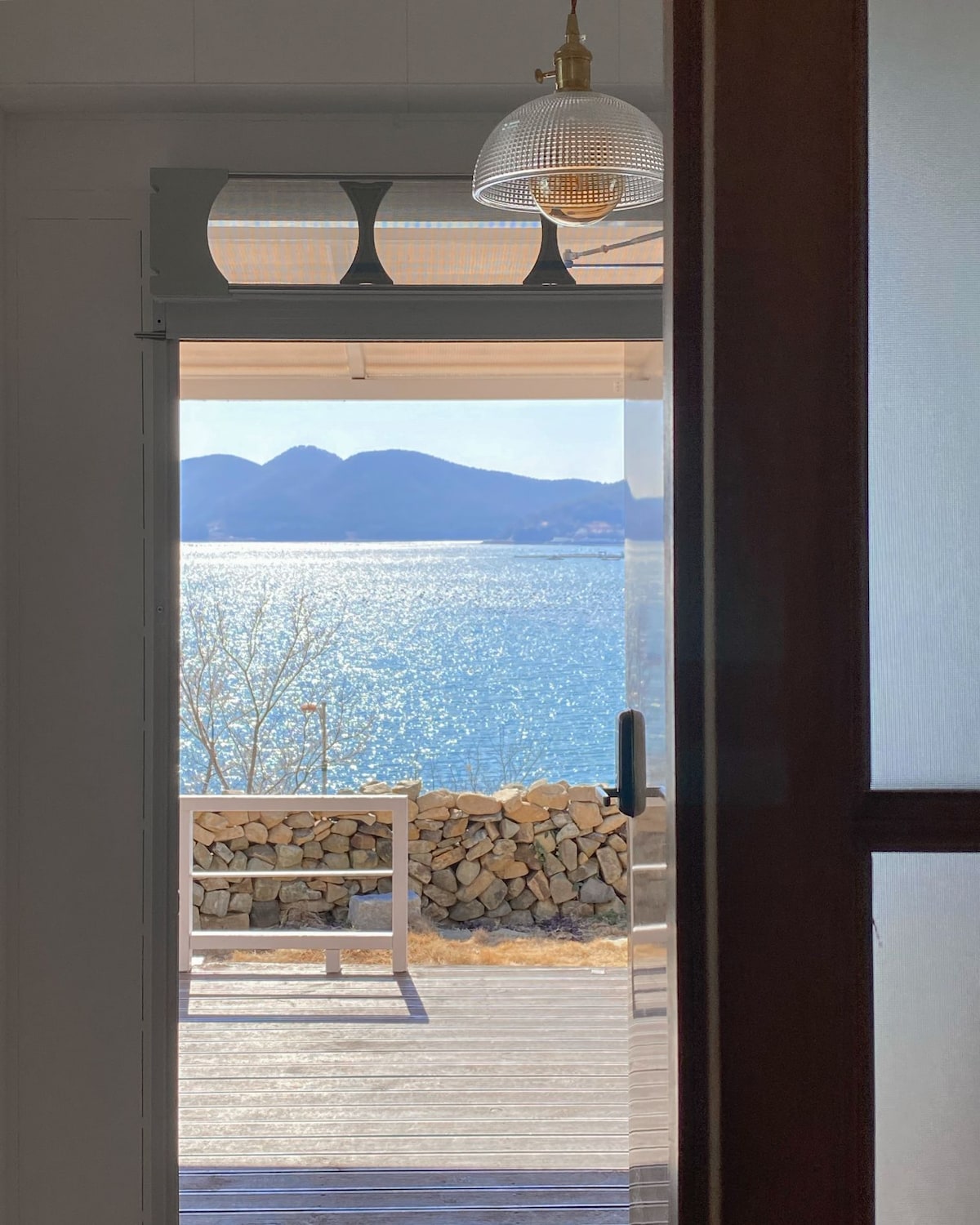 A doorway opens to reveal a scenic view of the shimmering blue sea and distant mountains. Sunlight reflects off the water, creating a bright atmosphere. A wooden bench is positioned on the outdoor deck, emphasizing the peaceful outdoor space.