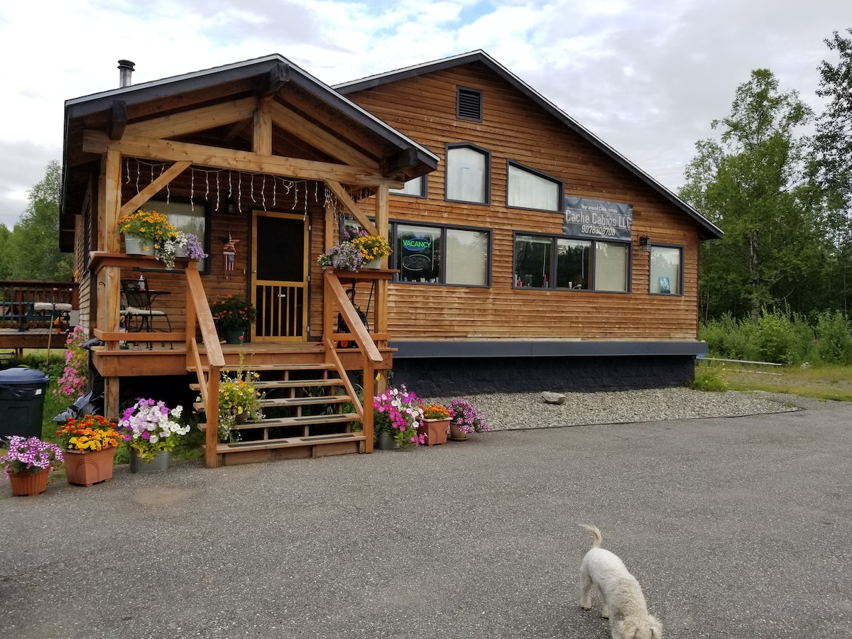 The exterior of a wooden cabin showcases a welcoming entrance with a spacious porch adorned with colorful flower pots. Large windows are visible along the front, offering natural light inside. A gravel parking area is seen in front, with a small white dog strolling nearby.