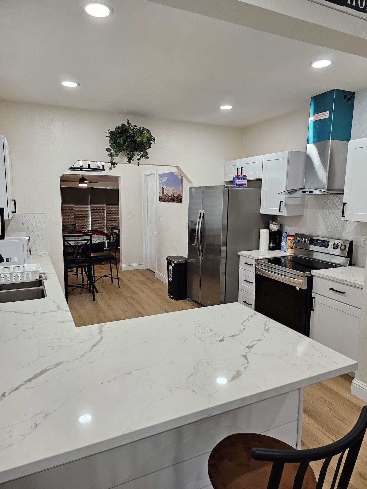 A modern kitchen is shown, featuring sleek white cabinetry and a spacious marble countertop. Stainless steel appliances, including a refrigerator and oven, are visible. Bright recessed lighting complements the neutral walls, and a small dining area can be seen in the background.