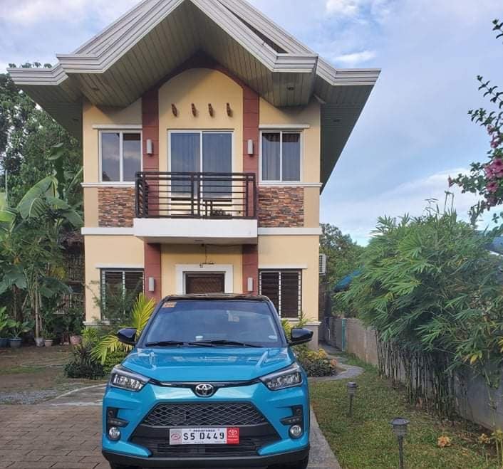 A modern two-story house is featured, showcasing a prominent facade with a combination of stone and painted walls. Large windows are present on both levels, providing natural light. A parked blue vehicle is seen in front, surrounded by well-maintained greenery and pathways.