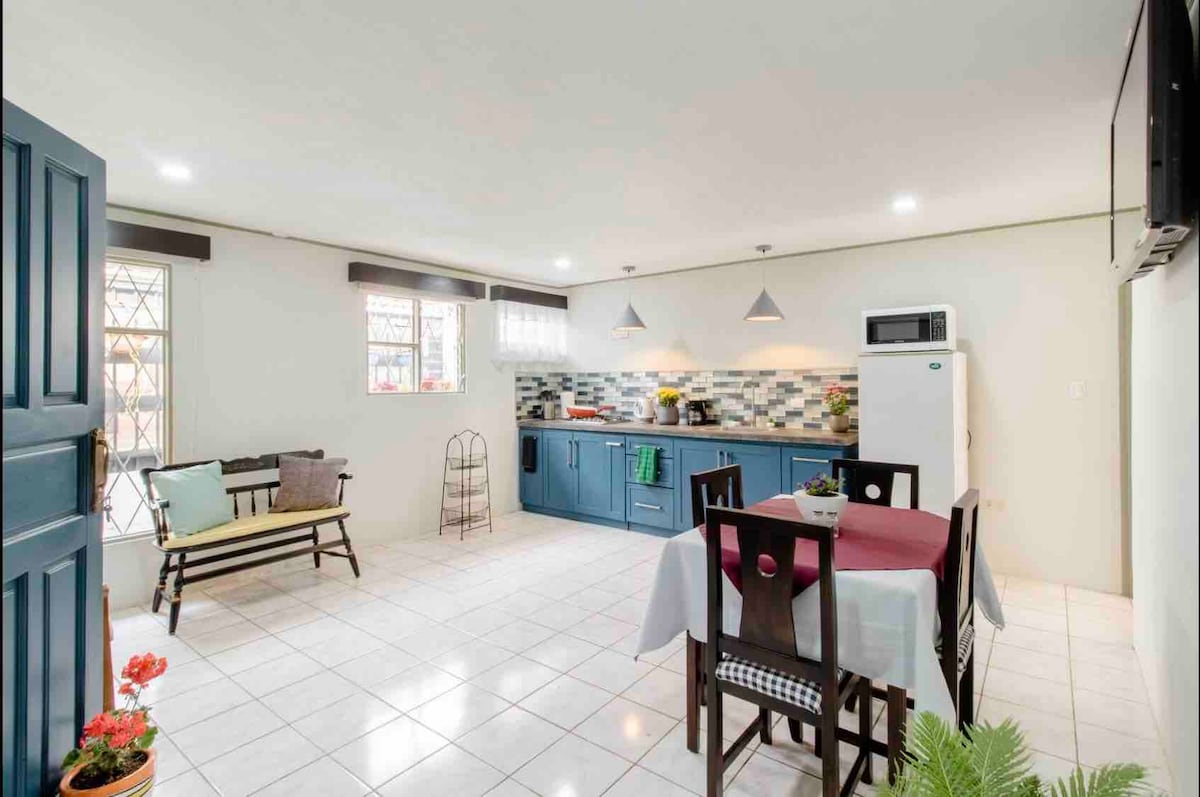 An open-plan kitchen and dining area is showcased, featuring blue cabinetry and a white countertop. A dining table is set for four, while a small seating area includes a bench. Large windows allow natural light to fill the space, creating a bright atmosphere.