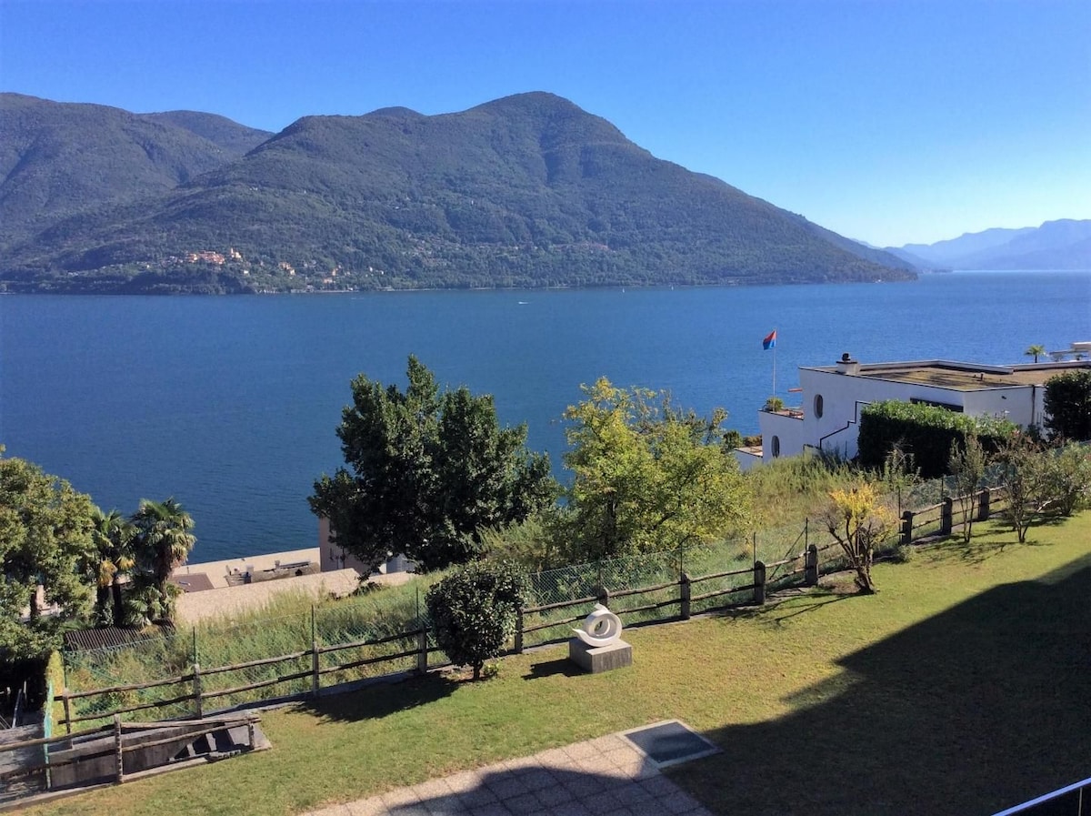 A scenic view of a tranquil lake surrounded by lush, green mountains under a clear blue sky. The landscape features a grassy area with shrubs, providing a serene backdrop for relaxation. The calm water reflects the natural beauty of the surroundings.