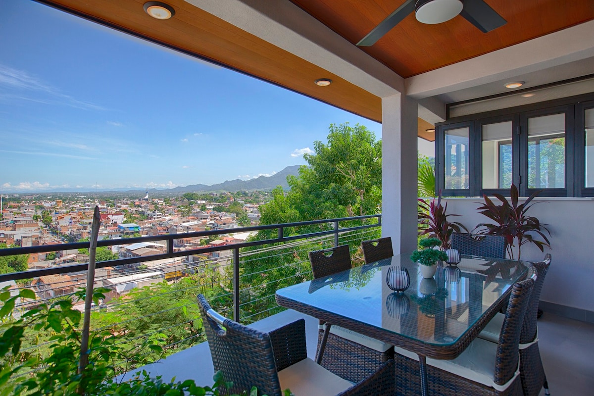 An outdoor dining area features a modern table with several chairs, surrounded by vibrant green plants. Large windows provide a panoramic view of the surrounding city and mountains, with clear skies enhancing the tranquil ambiance of the space.