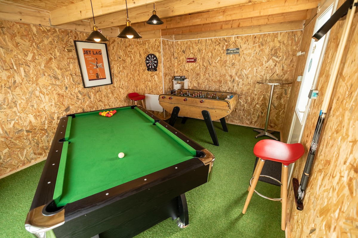 A game room is equipped with a pool table, foosball table, and dartboard, all set against a backdrop of wooden paneling. Green carpeting adds a cozy feel, while pendant lights provide ample illumination for evening play.