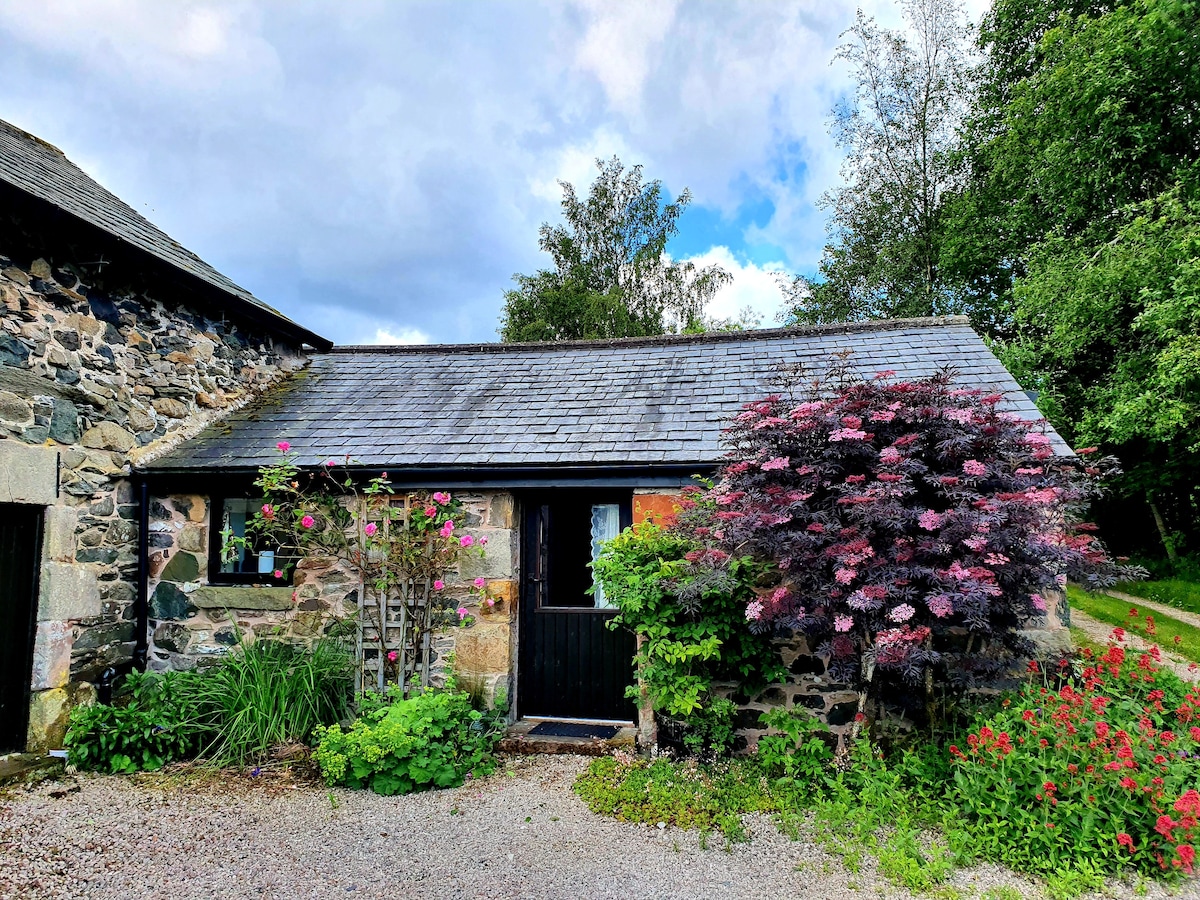 The exterior of a charming stone cottage is framed by lush greenery and colorful flowering bushes. A modest gravel path leads to the black door, which is adorned with a window featuring curtains. The roof is lined with slate tiles, complementing the natural surroundings.