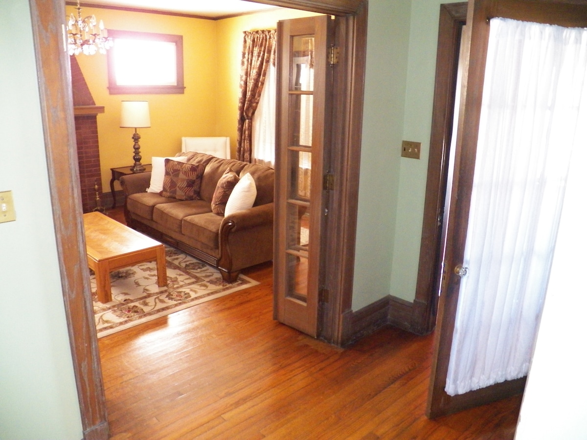 A living room area is viewed from an entryway, featuring a cozy brown sofa adorned with decorative pillows. A wooden coffee table sits on a patterned rug, and a fireplace adds character to the space. Natural light floods in from a nearby window, complemented by warm wall tones.