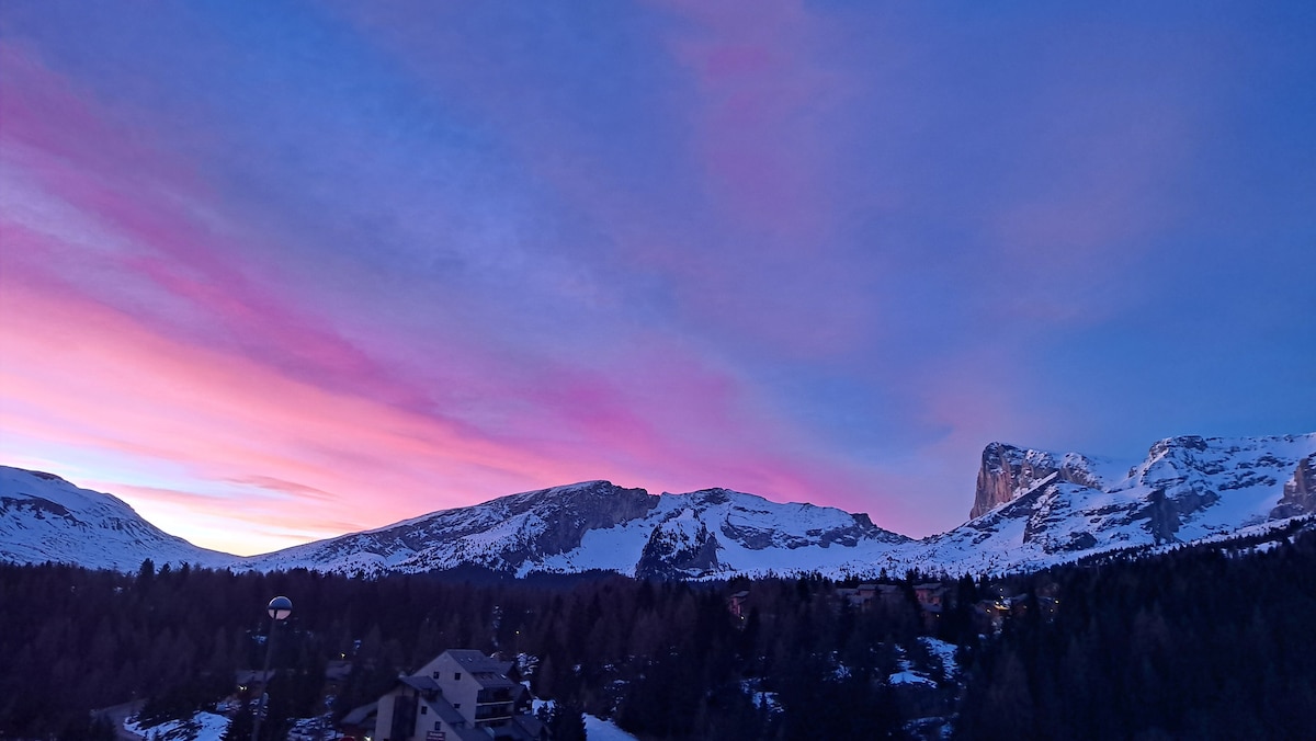 A panoramic view of a mountainous landscape features a breathtaking sunset displaying shades of pink and purple in the sky. Snow-covered peaks stand majestically against the vibrant colors, while nestled among the trees, buildings of the resort are visible at the base of the mountains.