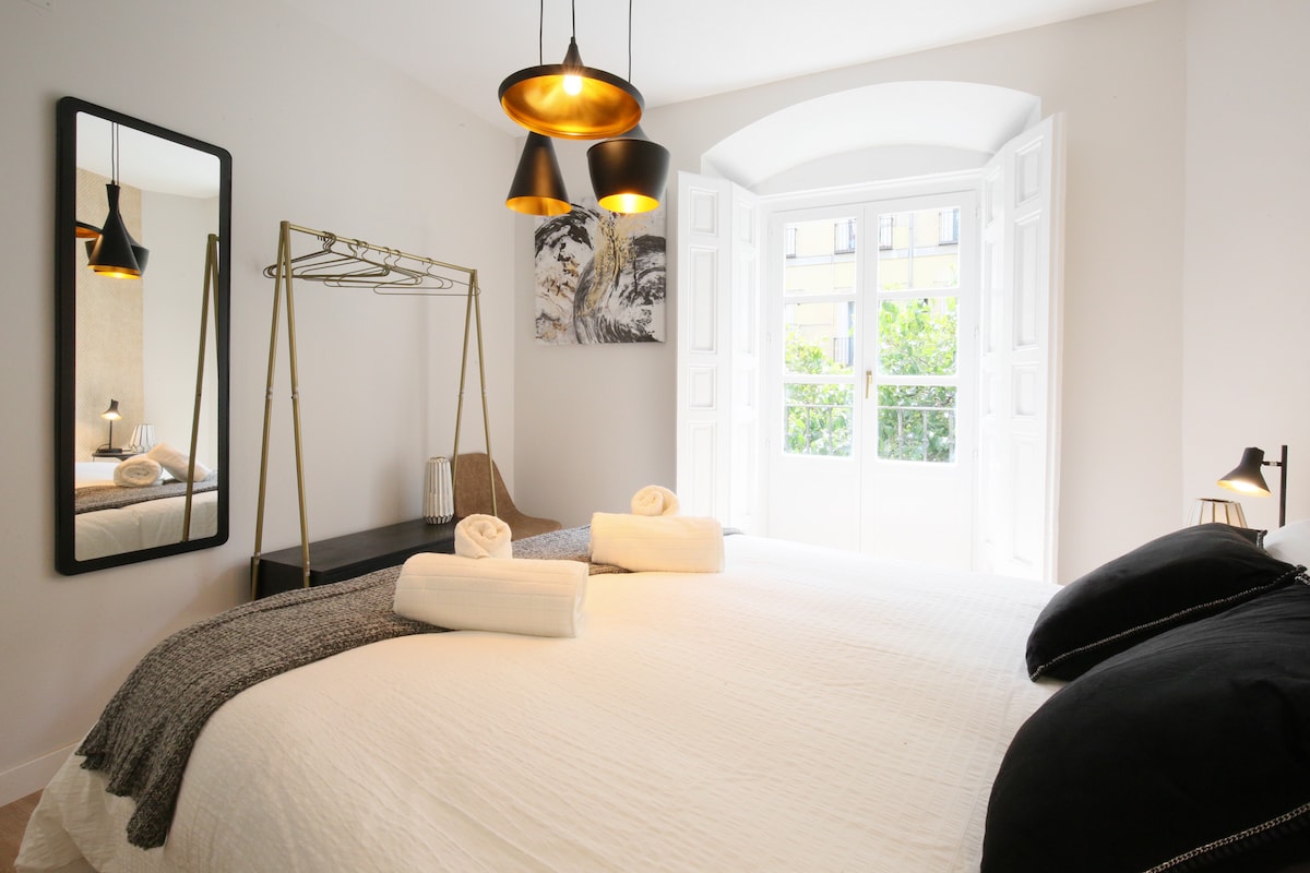 A well-lit bedroom features a spacious bed arranged with fresh linens and neatly rolled towels. A decorative mirror reflects the light from large windows, which open to greenery outside. Stylish pendant lights hang overhead, complementing the contemporary decor.