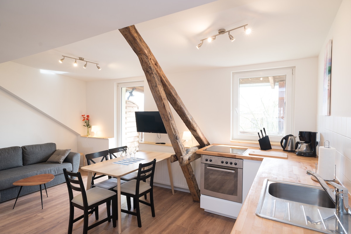 A modern kitchen area features wooden countertops and stainless steel appliances. A dining table with four chairs is positioned nearby. Light enters through two windows, illuminating a cozy living space with a gray sofa and a television mounted on the wall.