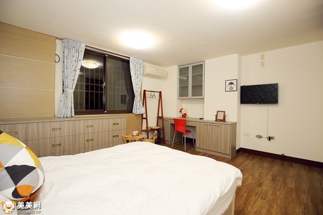 A spacious guest room is presented, featuring a large bed with white linens and a colorful accent pillow. A small desk with a red chair and a television is visible, along with a wardrobe and large window dressed with light curtains.