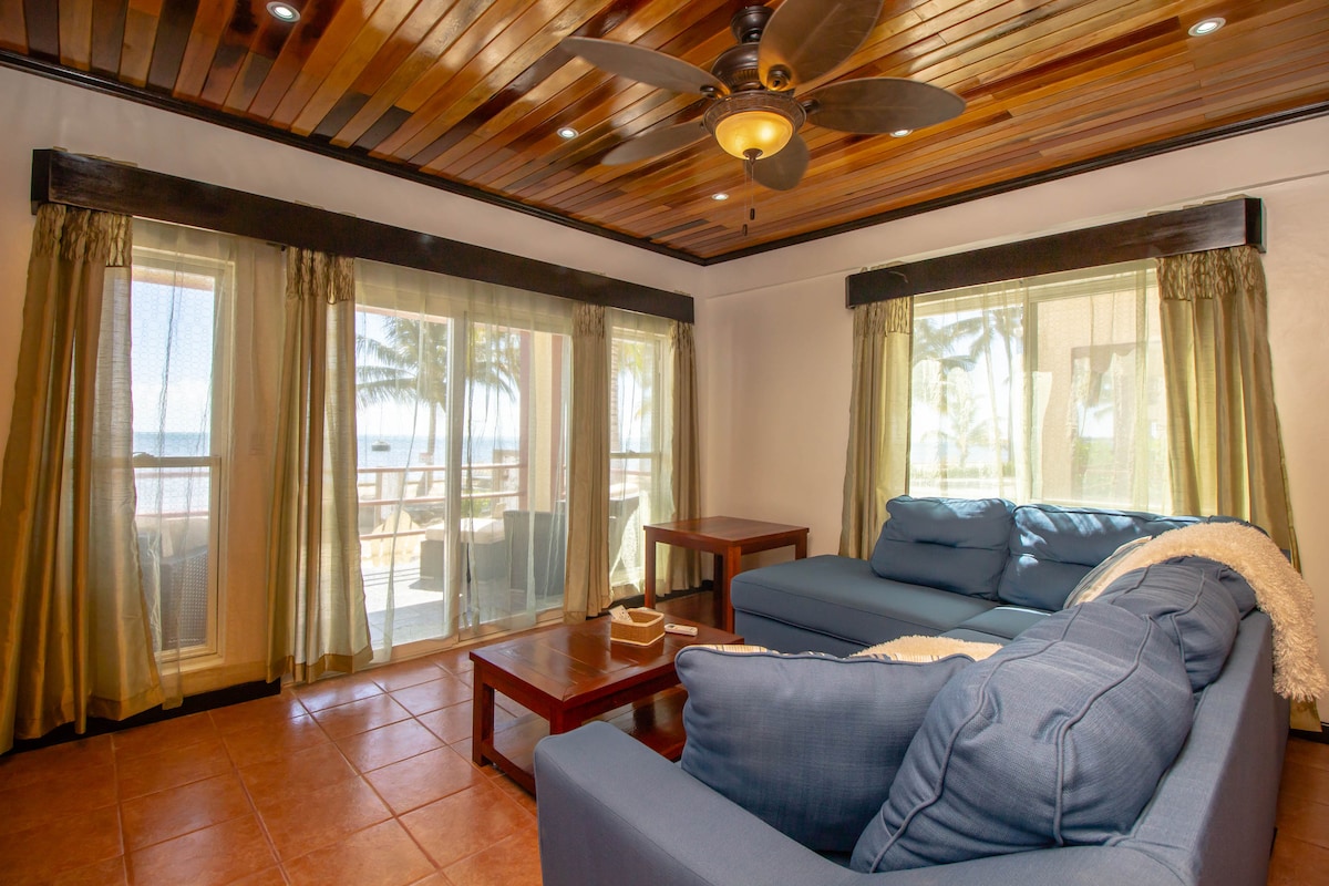 A comfortable living area features two blue sofas positioned around a wooden coffee table. Natural light floods through the large glass doors, which lead to a balcony adorned with palm trees. A ceiling fan with wooden blades enhances the airy ambiance.