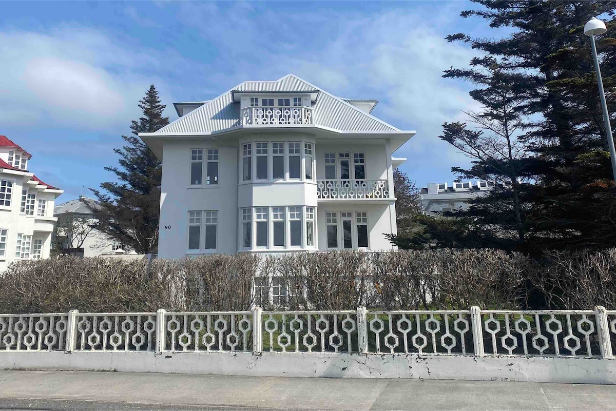 A spacious white building is framed by neatly trimmed hedges and a low decorative fence. Large windows are featured on the façade, allowing ample natural light. The structure's design includes gabled roofs and balconies, set against a clear blue sky.