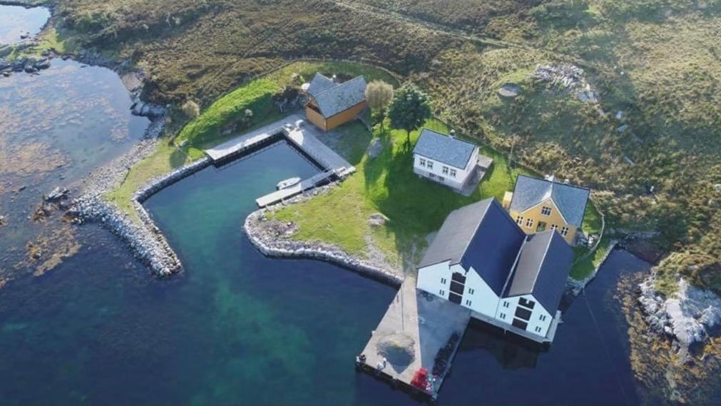 An aerial view of a small island reveals several buildings and a boat dock set against a backdrop of clear water and grassy terrain. The structures are varied in color, with a mix of traditional styles, and the surrounding landscape is dotted with rocks and patches of green vegetation.