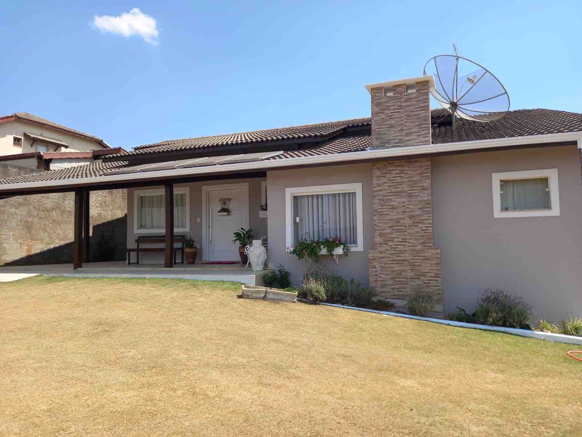 A single-story house features a combination of stone and stucco facades, with a covered porch at the entrance. Lush green grass frames the property, while potted plants add a touch of color. A satellite dish is visible on the roof, complemented by a bright blue sky.