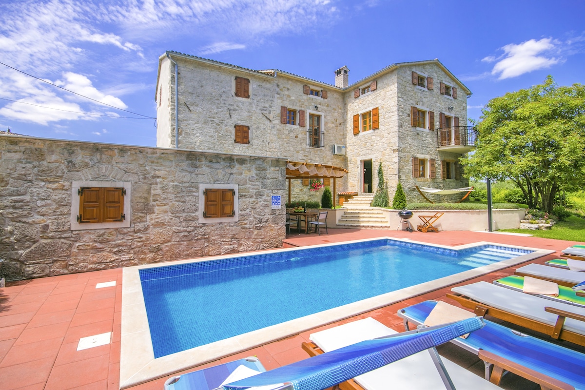 A classic Istrian villa constructed from stone is framed by lush greenery. A blue swimming pool is situated in the foreground, surrounded by sun loungers. The building features multiple balconies with wooden window shutters, complemented by a shaded outdoor dining area.
