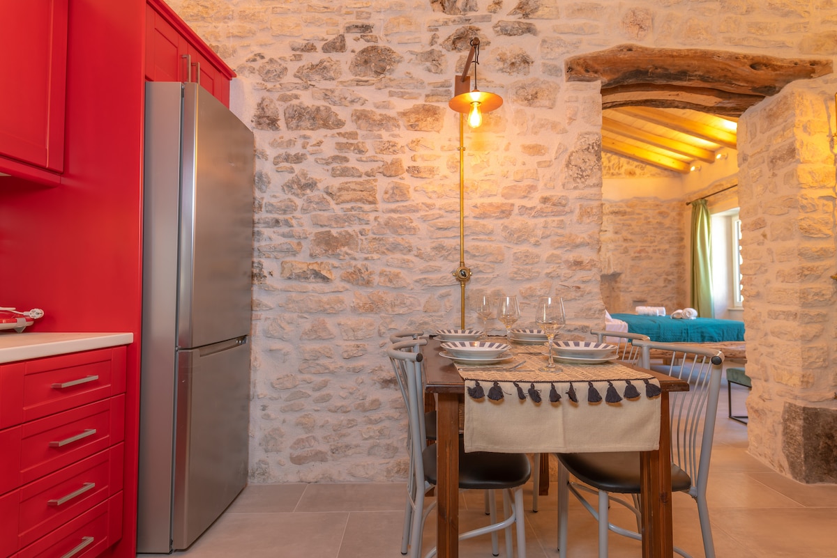 A dining area features a rustic wooden table set with dishes and glasses. Surrounding chairs are arranged neatly. Red cabinetry contrasts with the stone walls, while warm lighting adds a cozy ambiance. A glimpse of a blue-themed sleeping area is visible through the archway.