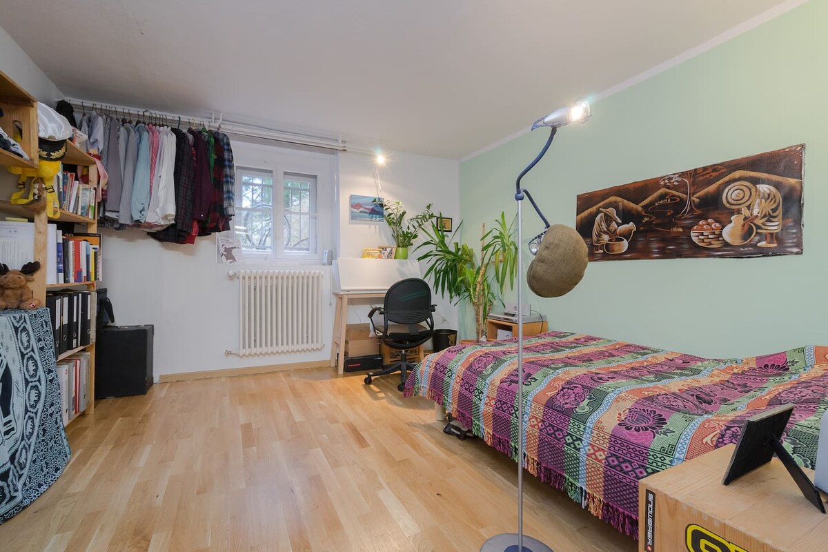 A private room is featured, showcasing a colorful patterned bedspread over a spacious bed. A desk and chair are positioned near a window, adjacent to a tall bookshelf filled with various items. Indoor plants add a touch of greenery, while warm light is provided by a floor lamp.