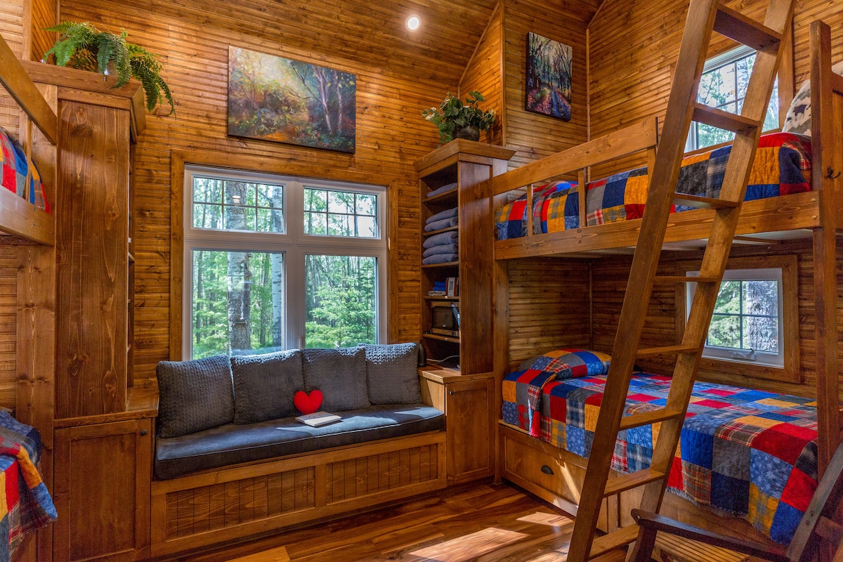 The cozy loft area features two wooden bunk beds draped in colorful quilts, alongside a built-in window seat adorned with a decorative heart pillow. Natural light streams in through multiple windows, highlighting the warm wood paneling and inviting cabin atmosphere.