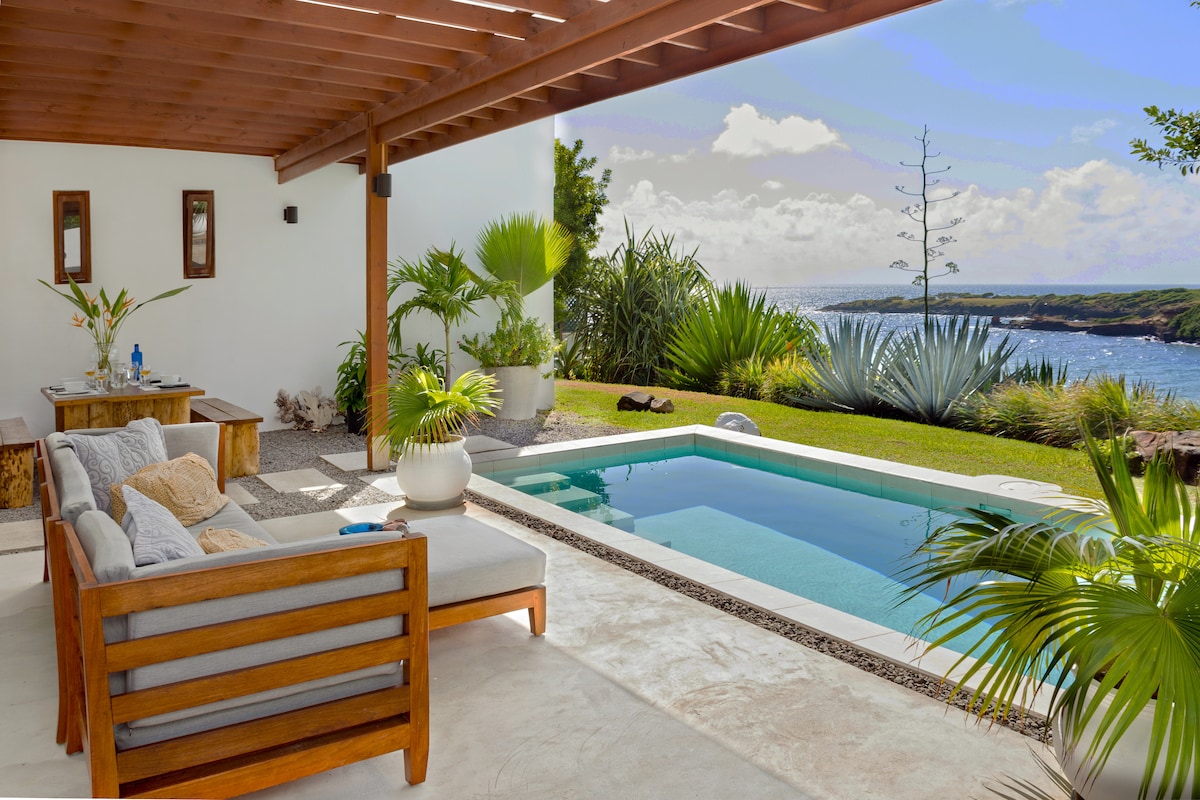 A private outdoor area features a seating arrangement with wooden furniture, providing a view of a relaxing swimming pool surrounded by tropical greenery. Coastal views are visible in the background, enhancing the secluded atmosphere of the space.