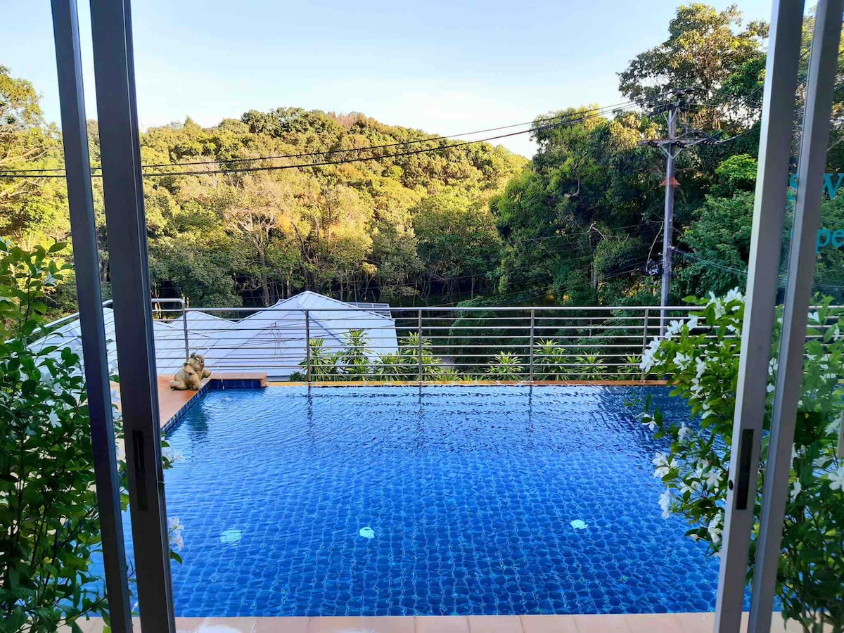 A serene view is offered from a glass-enclosed space, featuring a tranquil infinity pool that reflects the sky. Lush greenery surrounds the area, creating a peaceful atmosphere. The pool edge appears to merge seamlessly with the distant landscape of trees.